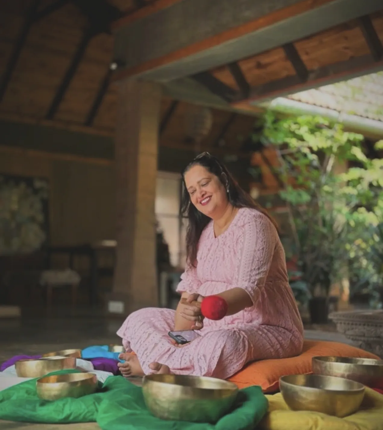 Sanggita Sharma seated among brass singing bowls on chakra-coloured cloths, smiling as she plays a bowl